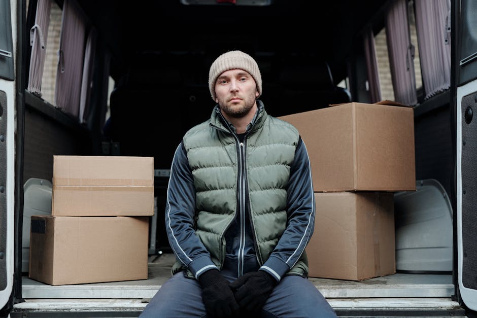A man with a serious expression, wearing a beige knit beanie, a green padded vest over a navy blue jacket, and black gloves, is seated on the edge of a black van with its rear doors open. Inside the v