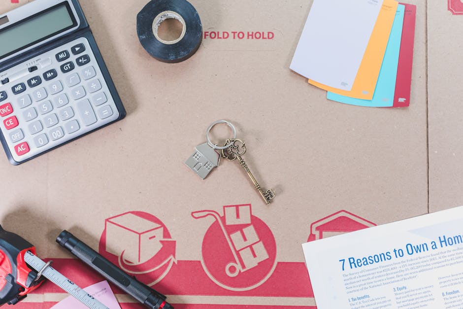 A close-up view of a cardboard box used for house removals, with various packing and office items arranged on top. Visible objects include a white calculator with grey and pink buttons, a roll of blac